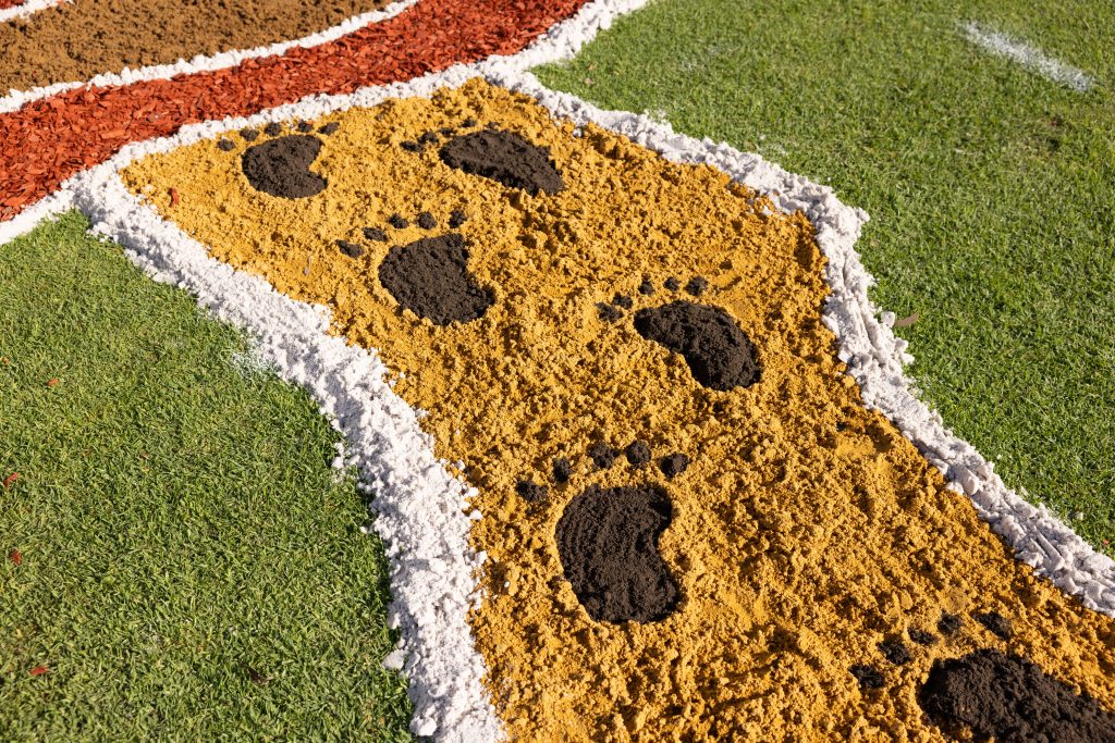 A close up of one part of the sand mural featuring a yellow sand path with black footprints, bordered by white sand, surrounded by green grass and areas of red and brown mulch